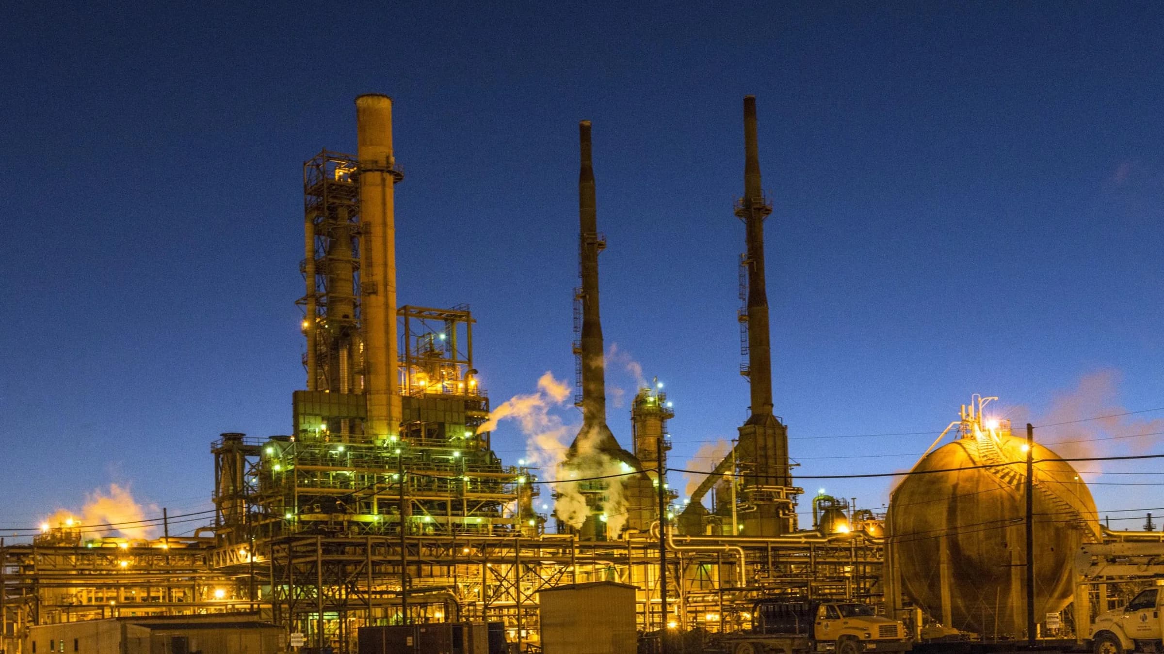 A Gulf Coast refinery at dusk, process units and flare stacks lit against a deep blue sky.