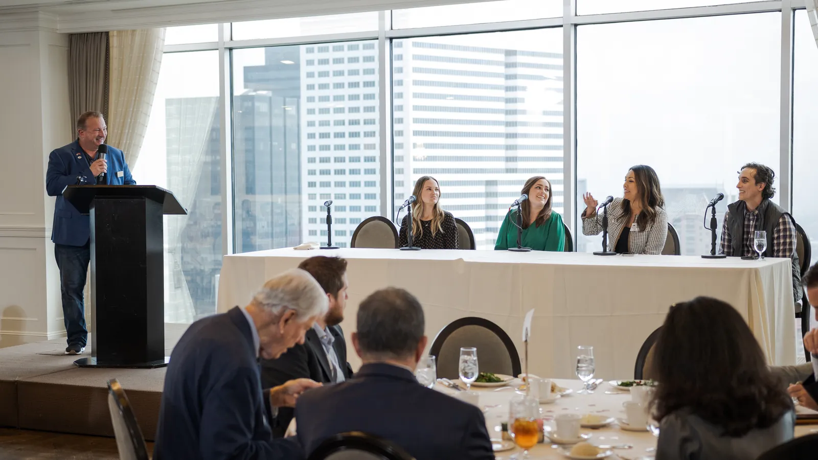 Mark LaCour at the podium hosting an oil and gas marketing panel, with Matthew Bertram and three other panelists seated at a table in front of the Houston skyline