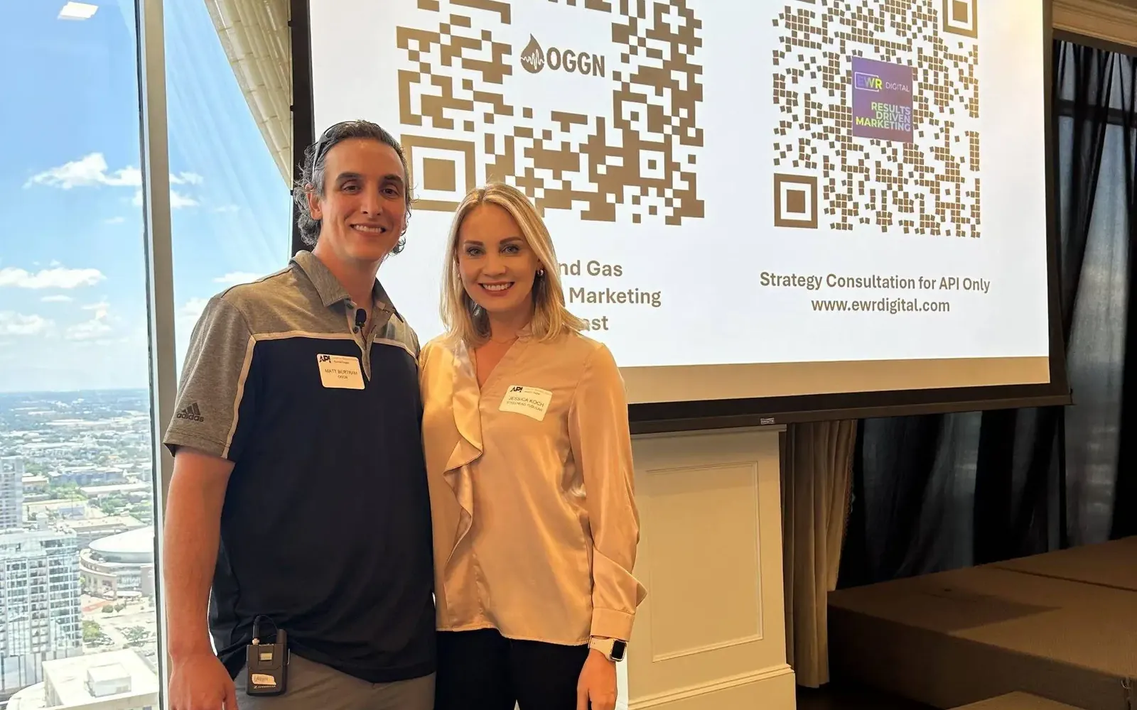Matthew Bertram with an attendee in front of a screen showing OGGN and EWR Digital QR codes, both wearing API Houston name badges, high above downtown Houston
